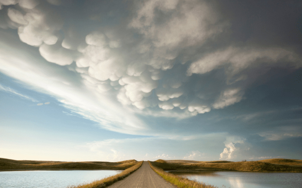 A rural dirt road stretches into the distance between two calm bodies of water, under a dramatic sky with large, puffy, textured clouds and a hint of sunlight breaking through.