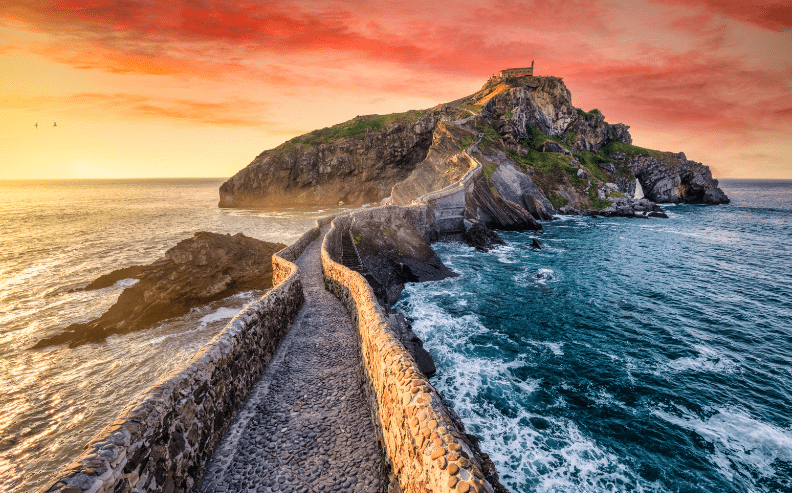 A winding stone walkway leads to a small chapel atop a rocky island surrounded by blue ocean waves, under a vibrant orange and pink sunset sky.