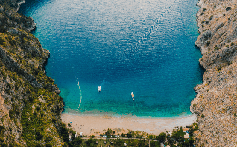 Aerial view of a secluded beach with clear turquoise water, surrounded by steep rocky cliffs. Two boats float near the shore, and waves gently lap against the sandy beach lined with greenery.