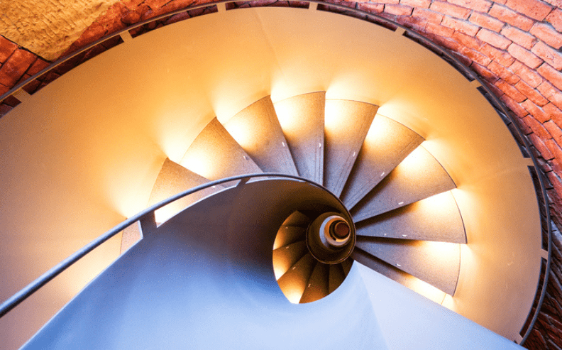 A top-down view of a spiral staircase with glowing lights along each step, surrounded by a curved brick wall. The railing winds smoothly down the center, creating a dynamic and symmetrical pattern.