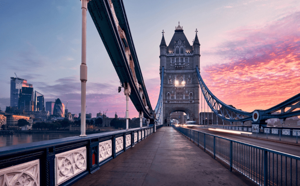 Tower Bridge in London at sunrise, with a colorful sky and modern city buildings in the background. The bridge appears empty, highlighting its detailed architecture and blue railings.
