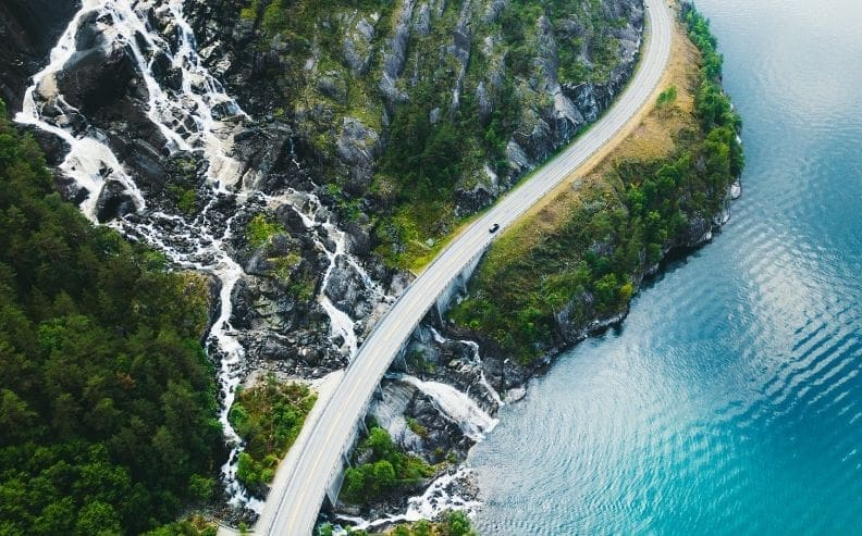 A winding road runs alongside a rocky cliff with cascading waterfalls on one side and a vibrant blue lake on the other, surrounded by lush green trees.