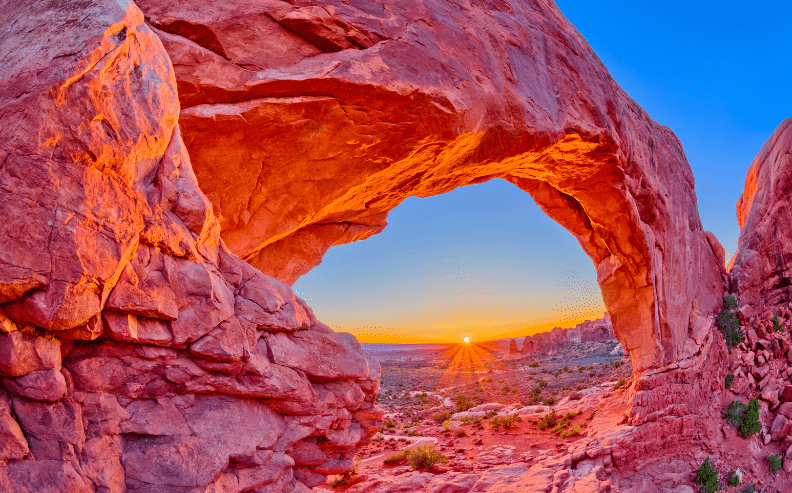 A vibrant sunrise viewed through a large natural rock arch in a desert landscape, with the sky transitioning from deep blue to orange and the rocky formation glowing with warm sunlight.