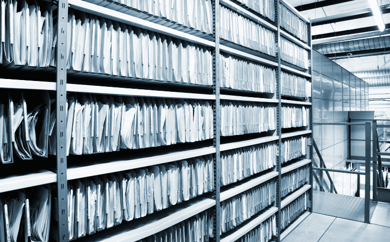 Rows of shelves filled with organized paper files and folders in an archive or records storage room, with a modern, industrial interior visible in the background.