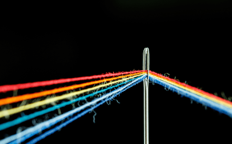 A close-up of a sewing needle with multiple colorful threads—red, orange, yellow, green, blue, and purple—running through its eye against a black background.