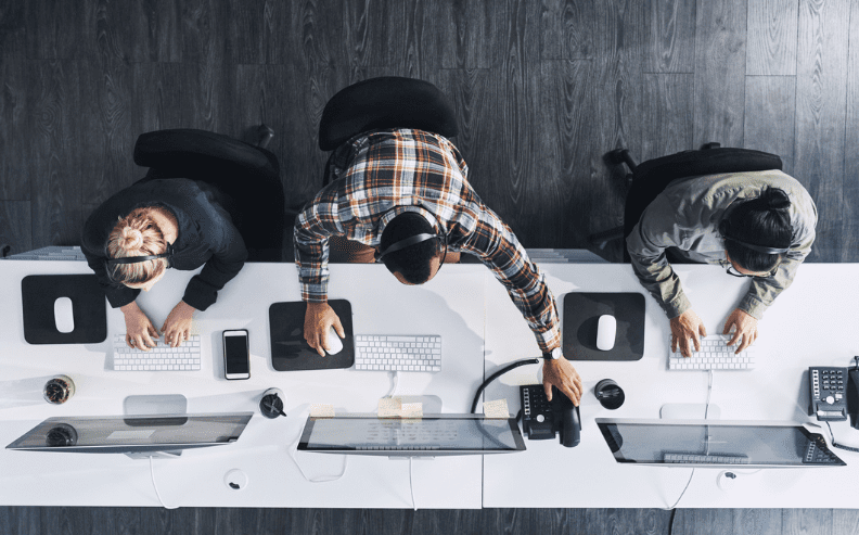 Aerial view of three people wearing headsets, sitting at a white desk with computers and keyboards, collaborating on M&A identity integration in an office environment with dark wooden flooring.