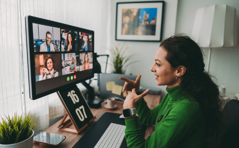 A woman in a green sweater sits at a desk, participating in a video call with six people displayed on her computer monitor, smiling and gesturing with her hands. Her workspace includes plants and a digital clock.