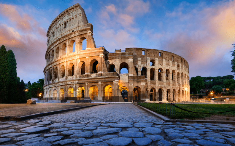 The Colosseum in Rome at sunset, illuminated with warm lights. The ancient stone amphitheater is partially ruined, with clouds in the sky and cobblestone path in the foreground. Greenery surrounds the structure.