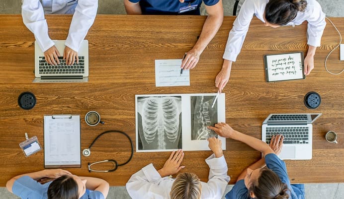 Five healthcare professionals sit around a wooden table with laptops, papers, and X-ray images, collaborating and discussing patient information. A stethoscope and coffee cups are also visible on the table.