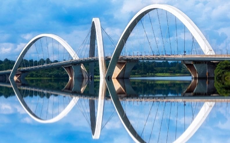 A modern bridge with three large, curved arches spans over a calm river, perfectly reflecting the structure and blue sky with clouds in the water below. Lush greenery lines the riverbanks.