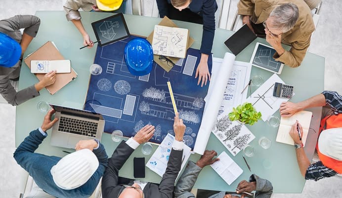 A group of people wearing hard hats and business attire sit around a table with blueprints, architectural plans, laptops, and notebooks, collaborating on a construction or design project.