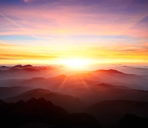 Sunrise over mountains with a glowing sun on the horizon, casting warm orange, yellow, and pink light across a misty landscape and soft clouds in the sky.