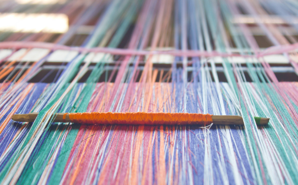 Close-up of colorful threads stretched on a loom, with an orange-wrapped shuttle placed among the threads, highlighting the weaving process and vibrant yarns.