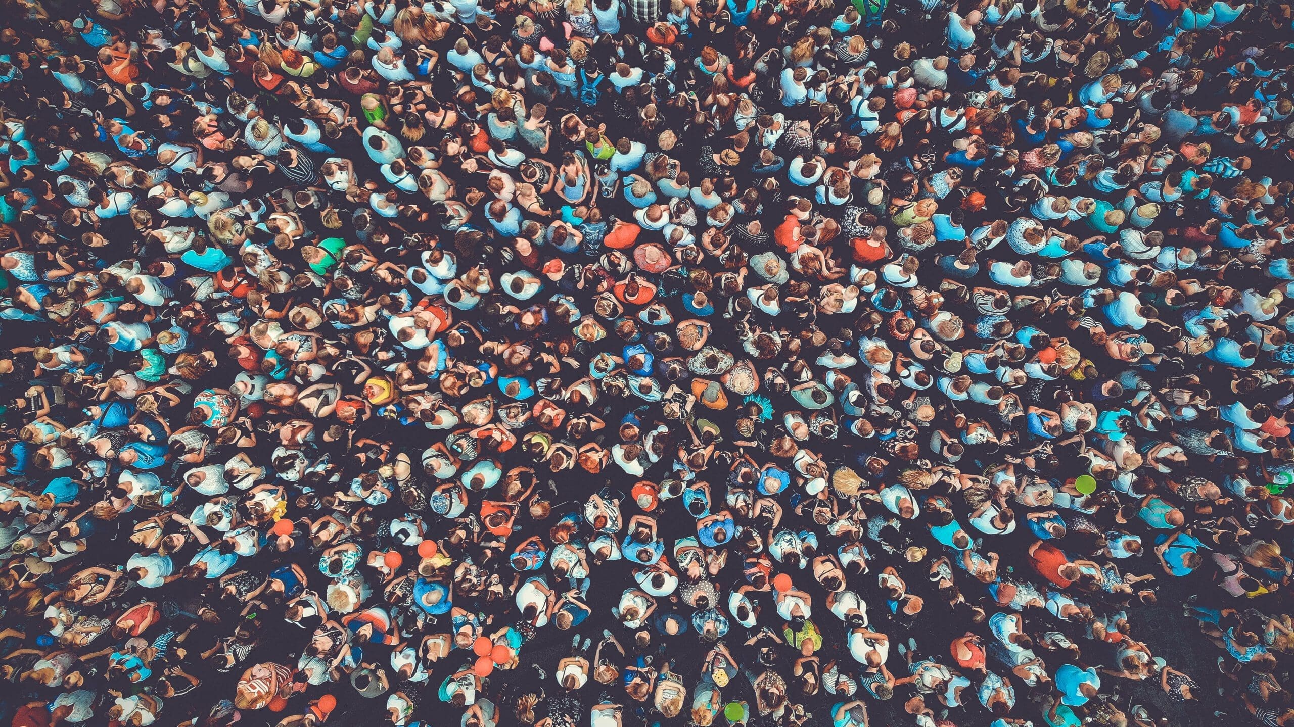 Aerial view of a large, dense crowd of people standing close together, wearing colorful clothing and hats, with some individuals looking up toward the camera.