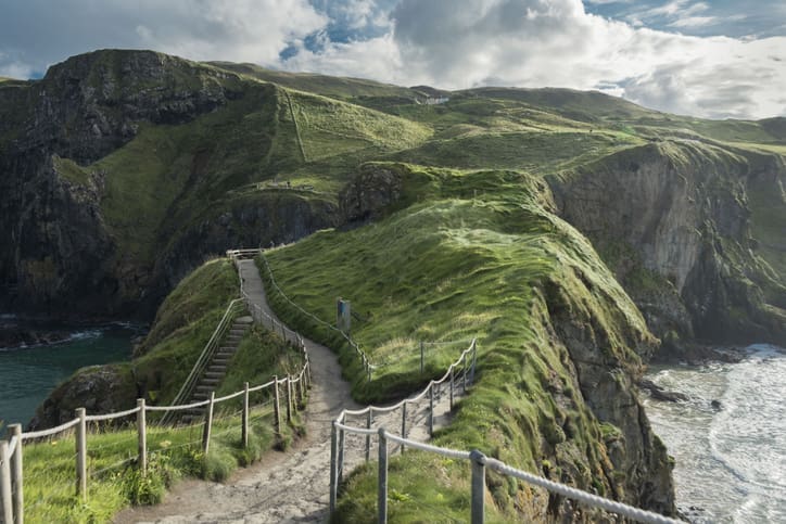 A narrow path with rope railings winds along a grassy, rugged cliff edge above the ocean, with steep cliffs and rolling green hills in the background under a partly cloudy sky.