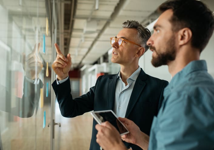 Two men standing by a glass wall with sticky notes, one pointing at the notes while the other holds a tablet, discussing ideas in a modern office setting.