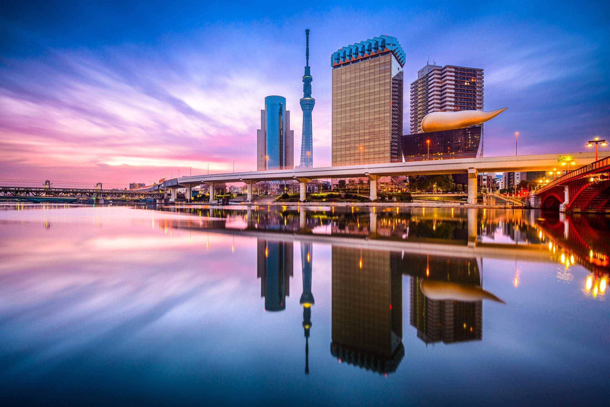 Tokyo skyline at sunset with modern buildings, including Tokyo Skytree and the golden Asahi Beer Hall, reflected in the calm waters of the Sumida River under a colorful sky.