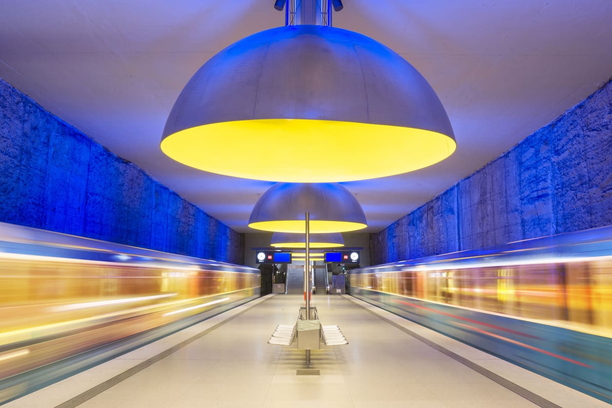 A brightly lit subway station with large yellow and gray dome lights, blue illuminated walls, and two trains passing by on either side, creating motion blur. Benches are placed in the center of the platform.