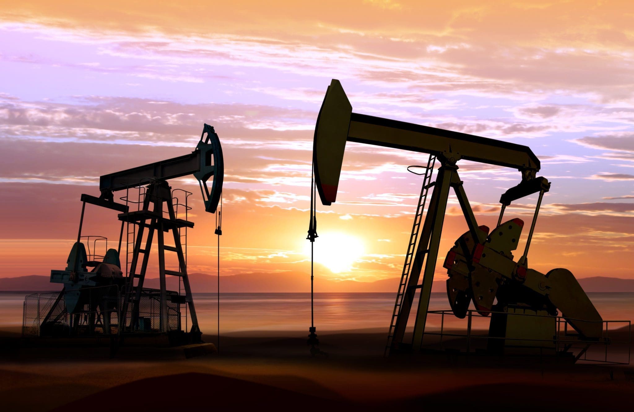 Two oil pumpjacks silhouette against a colorful sunset sky, with the sun low on the horizon and clouds scattered above, creating a dramatic and industrial landscape.