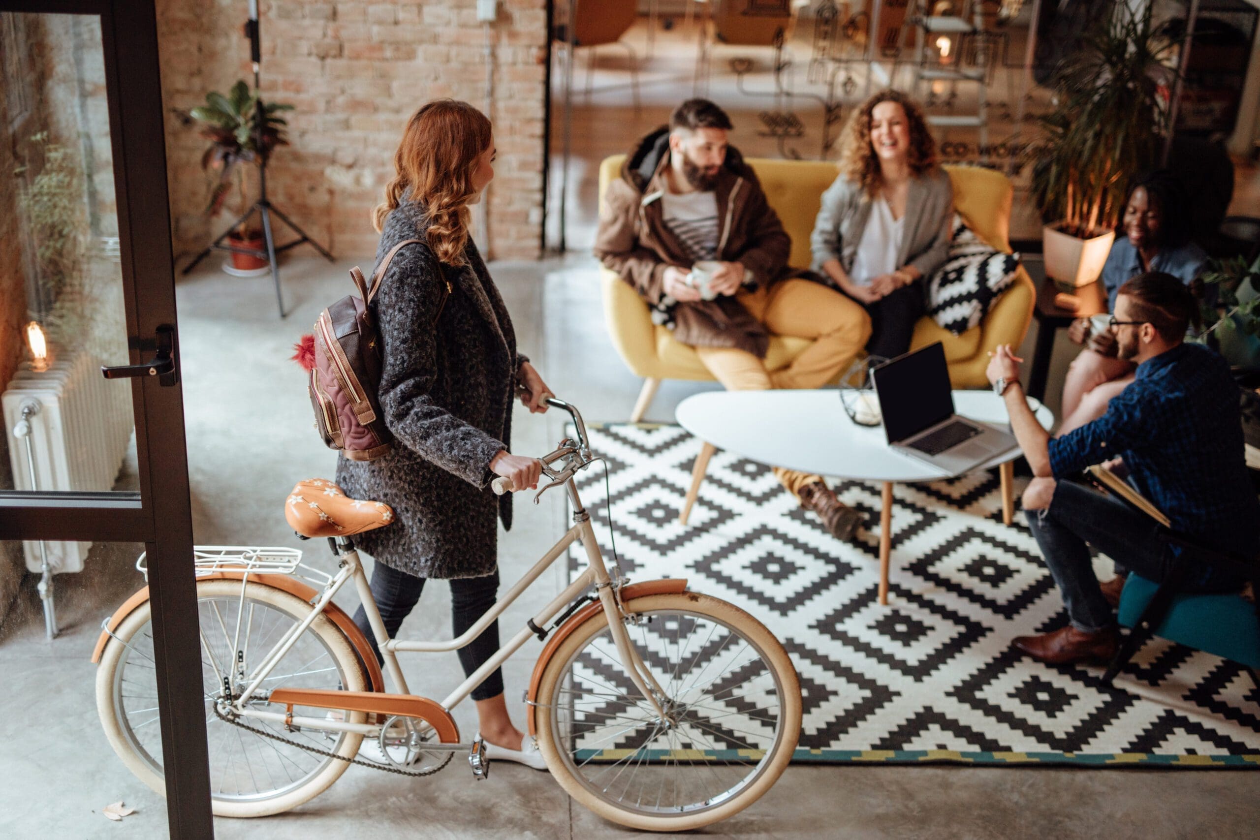 A woman with a backpack walks a bicycle into a stylish, modern office where four people sit on a couch and chairs around a white coffee table, talking and smiling. A laptop is open on the table, and plants decorate the room.