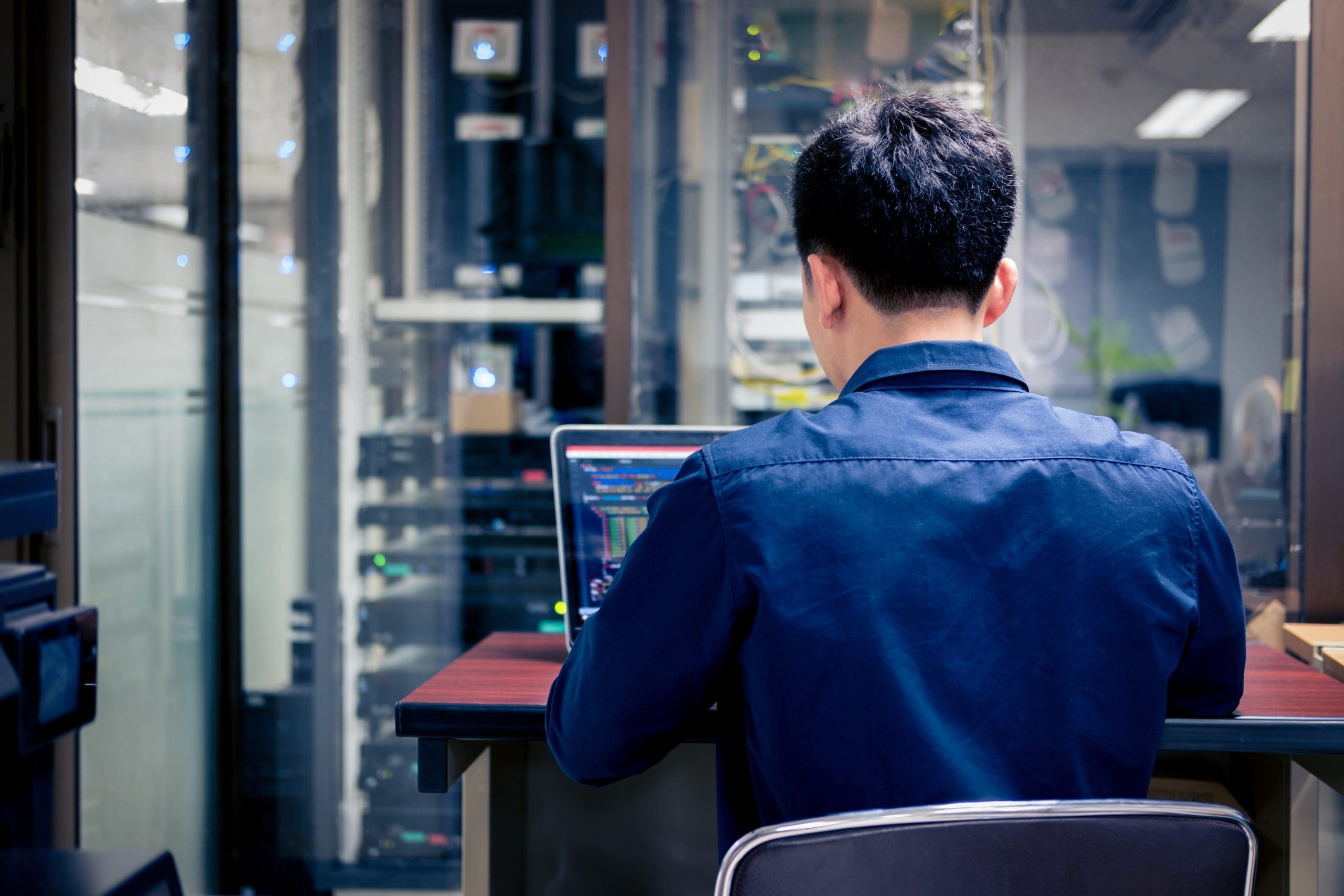 A person sits at a desk in front of a laptop, working in a server room filled with racks of computer servers and network equipment. The person is seen from behind, wearing a dark blue shirt.