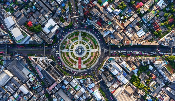 Aerial view of a large, circular city roundabout with roads radiating outward, surrounded by densely packed buildings and colorful rooftops. Traffic flows along the streets encircling the central landscaped area.